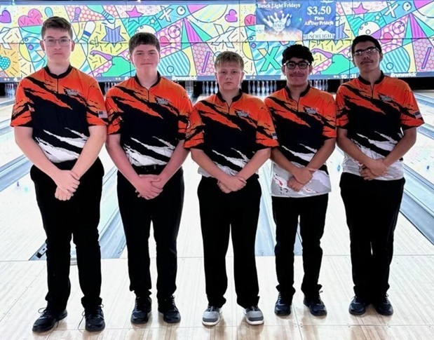 Five male teens in orange, black and white bowling shirts pose for a photo at a bowling alley.