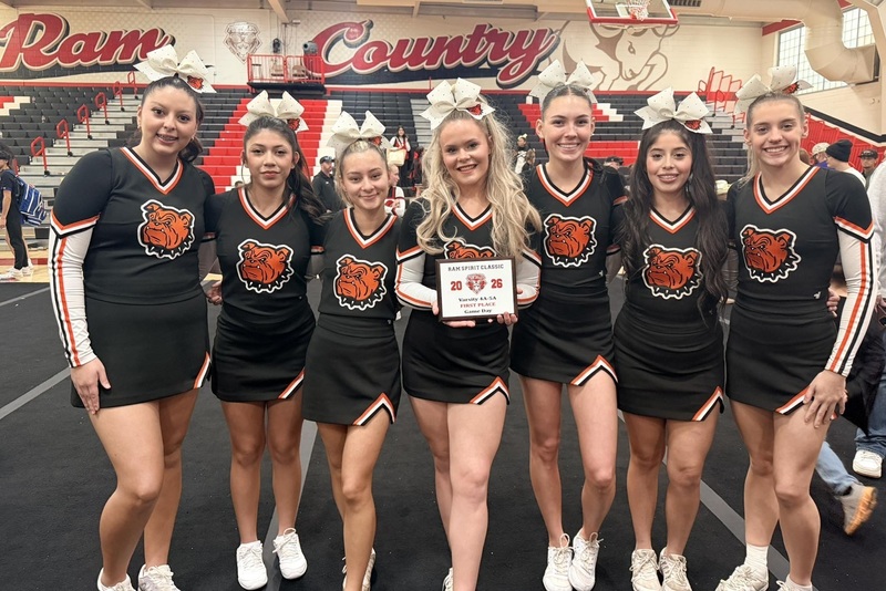 Seven female teens in black cheerleading outfits with orange Bulldog heads and large white bows in their hair pose for a photo. The center teen holds a plaque.