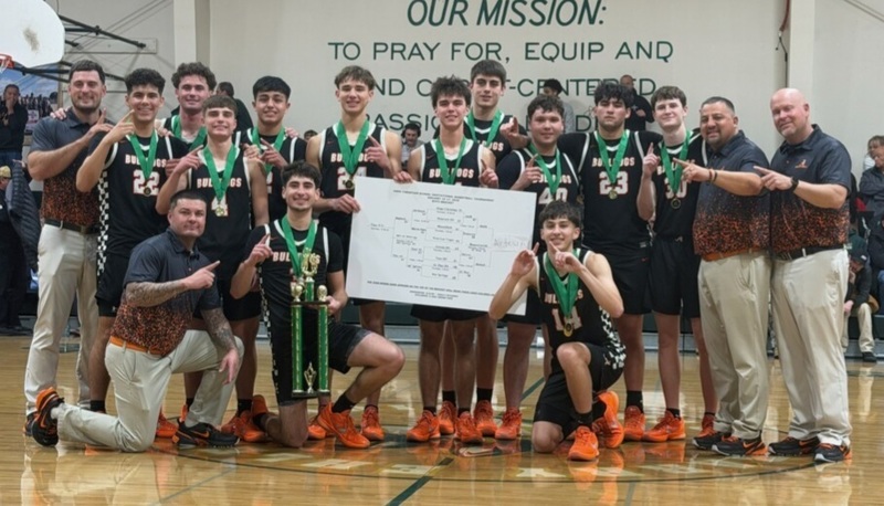 A large group of male teams in black and white basketball uniforms poses with a trophy and large bracket alongside four male coaches. All team members wear a medal on a green ribbon around their necks.