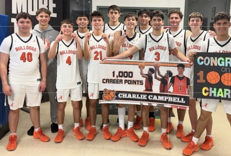A group of teen boys in white and orange basketball uniforms and orange shoes pose for a photo with a teammate holding a banner reading: "1,000 Career Points: Charlie Campbell".