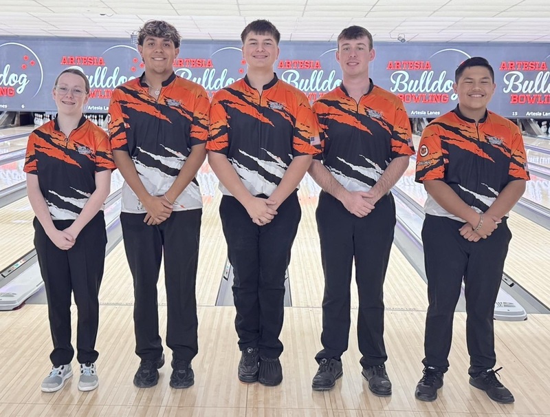 One female and four male teens in orange, black and white bowling shirts pose for a photo in front of bowling lanes.