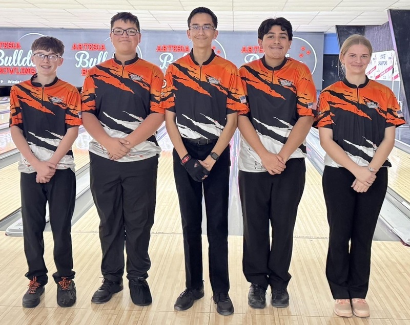 One female and four male teens in orange, black and white bowling shirts pose for a photo in front of bowling lanes.