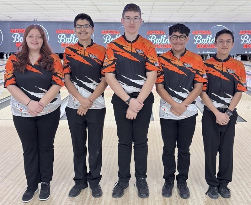 One female and four male teens in orange, black and white bowling shirts pose for a photo in front of bowling lanes.