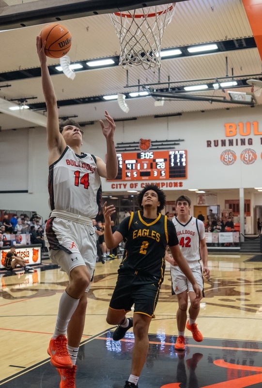 A teen boy in a white and orange basketball uniform goes up under the basket to lay the ball in with his right hand as an opponent in black and gold looks on.