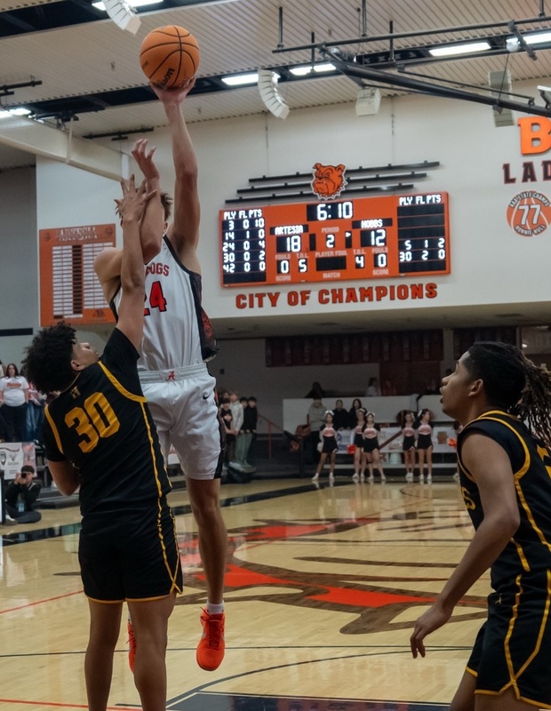 A teen boy in a white and orange basketball uniform jumps in the lane to put the ball up with his left hand as an opponent in black and gold raises a hand to his face to defend.