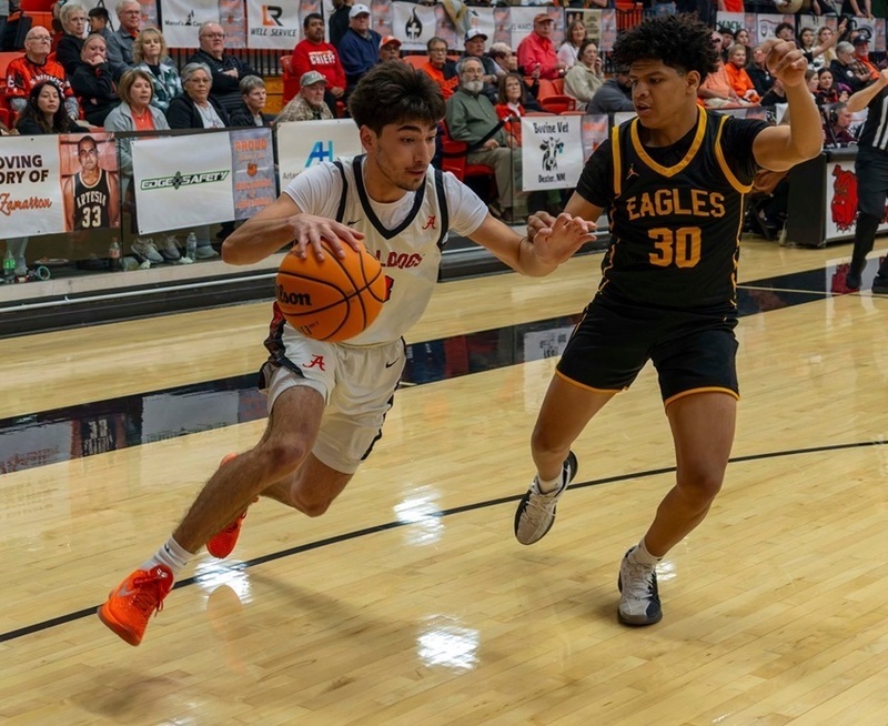 A teen boy in a white and orange basketball uniform dribbles hard around an opponent in black and gold as fans look on from courtside seats.