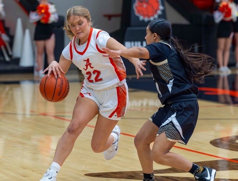 A teen girl in a white and orange basketball uniform dribbles around an opponent in black and grey while holding out her left hand to put space between herself and the defender.