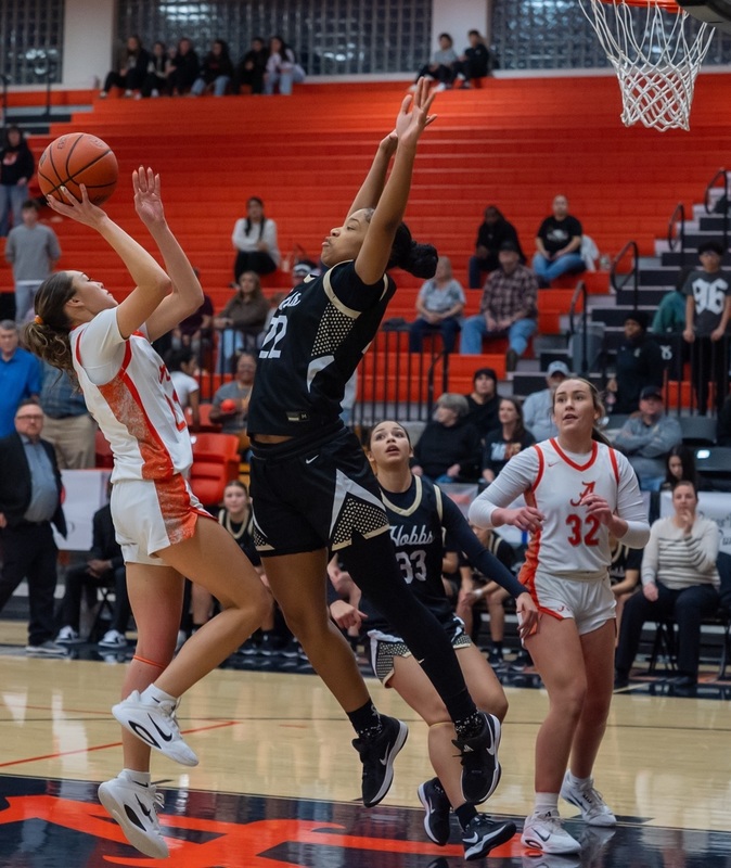 A teen girl in a white and orange basketball uniform jumps as she prepares to release the ball from her right hand as an opponent in black and grey leaps in front of her with both arms raised above her head.