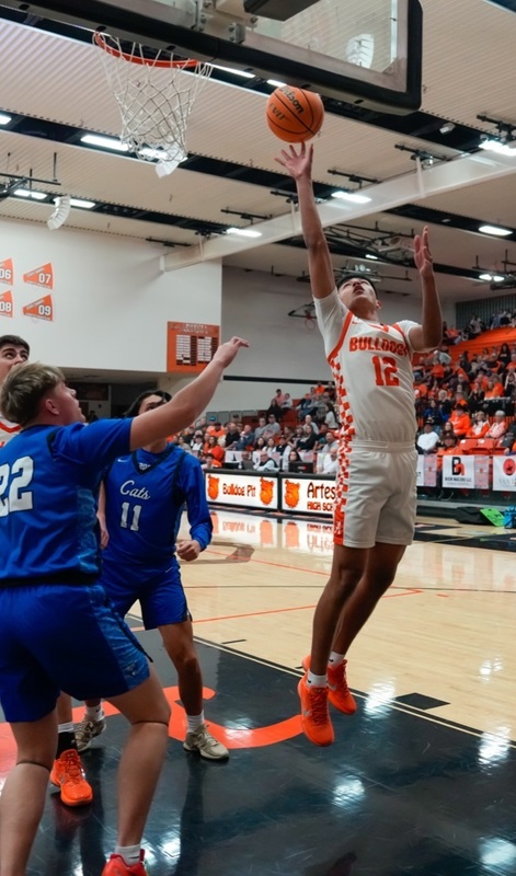 A teen boy in a white and orange basketball uniform lays the ball off the glass with hi right hand as defenders in blue look on.