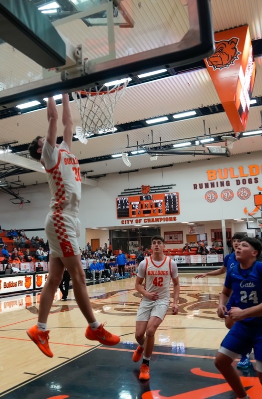 A teen boy in a white and orange basketball uniform dunks the ball as a teammate and two defenders in blue look on.