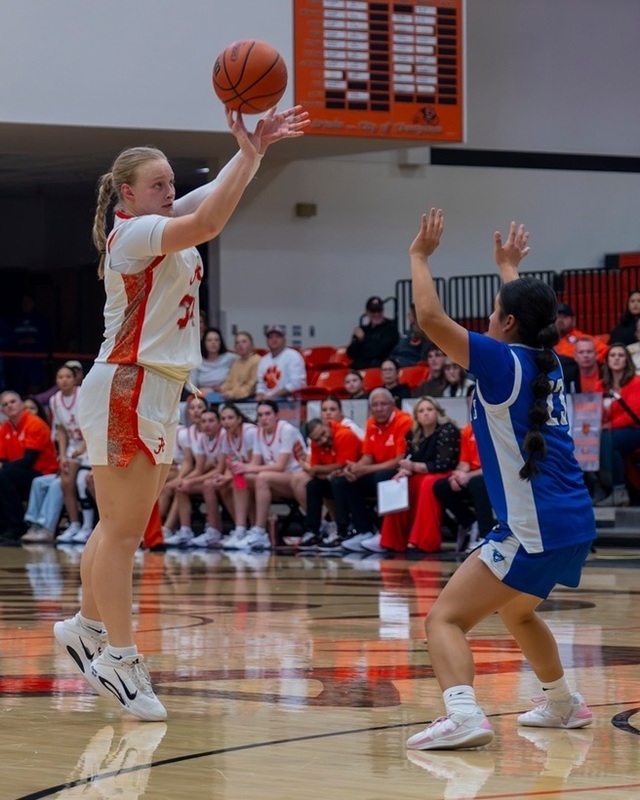 A teen girl in a white and orange basketball uniform goes up on her toes to shoot a three as an opponent in blue and white defends.