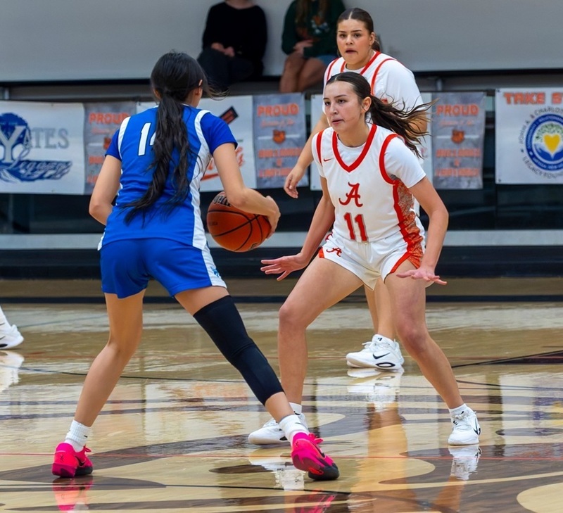 A teen girl in a white and orange basketball uniform is shown in a defense stance facing an opponent in blue and white.