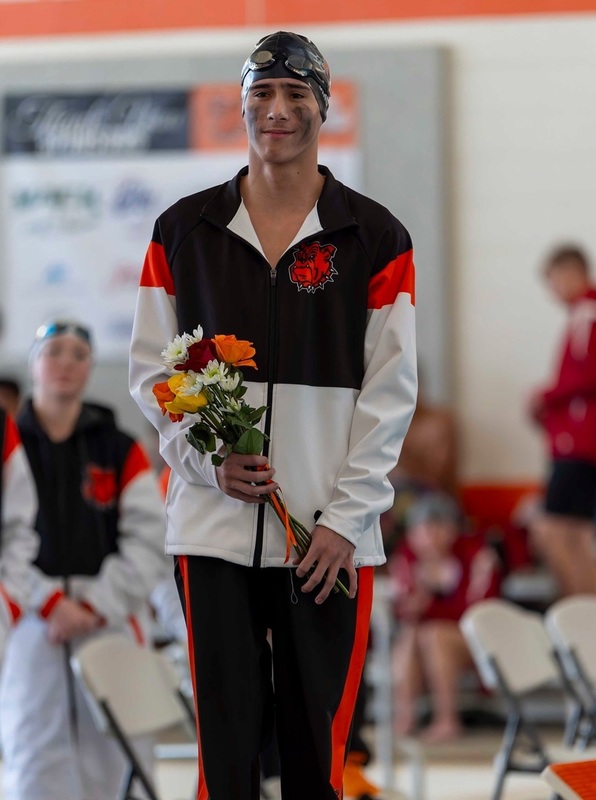 A teen boy in white, orange and black swim warm-ups and a black swim cap holds a small bouquet of orange and white flowers.