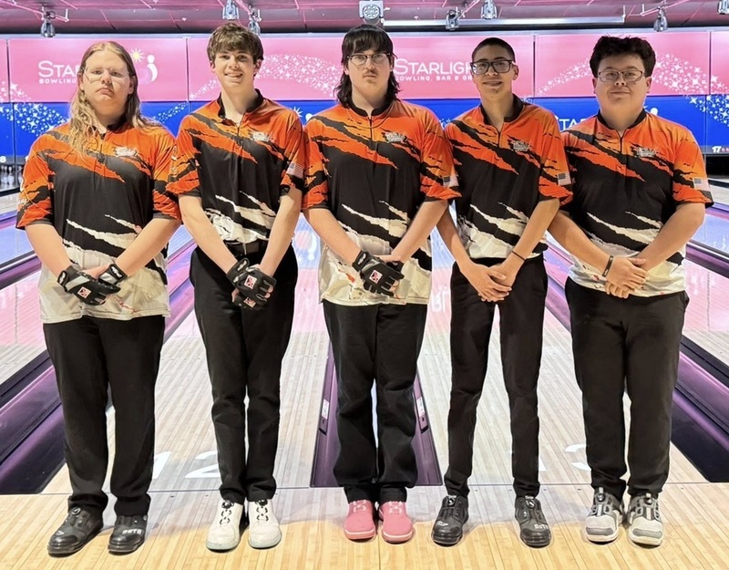 Five male teens in orange, black and white bowling shirts pose for a photo in front of bowling lanes.