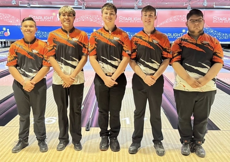 Five male teens in orange, black and white bowling shirts pose for a photo in front of bowling lanes.