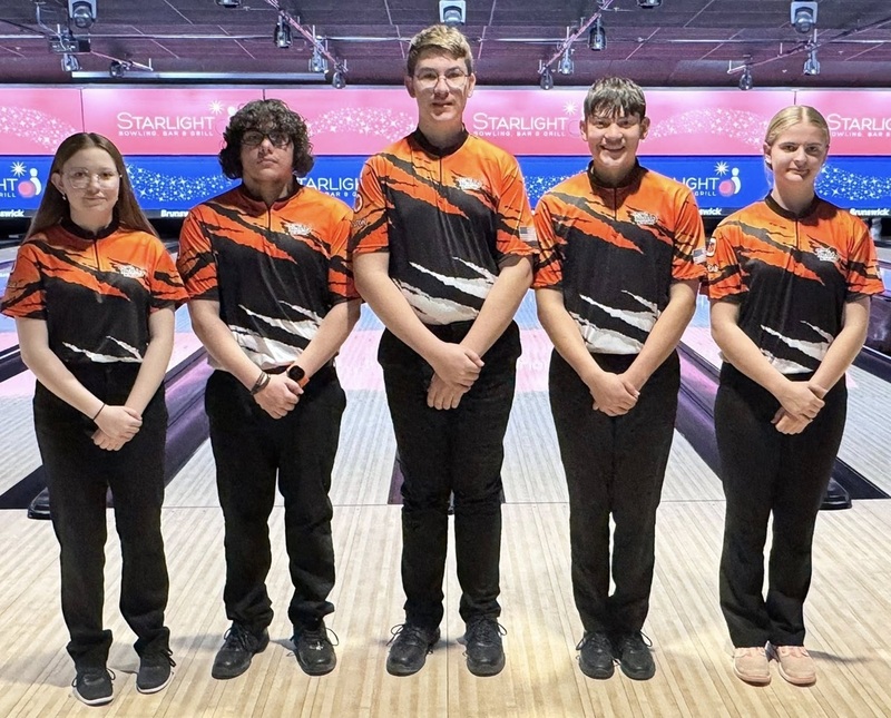 Two female and three male teens in orange, black and white bowling shirts pose for a photo in front of bowling lanes.