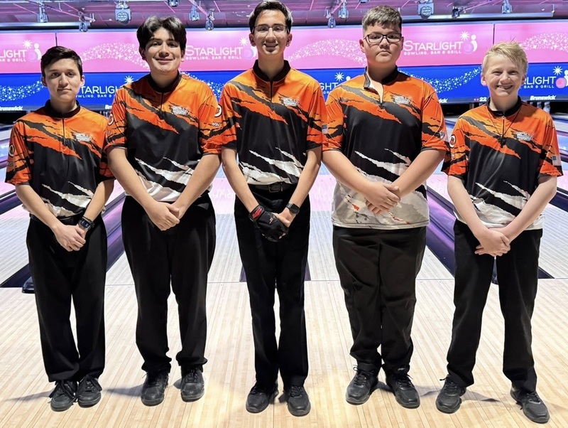 Five male teens in orange, black and white bowling shirts pose for a photo in front of bowling lanes.