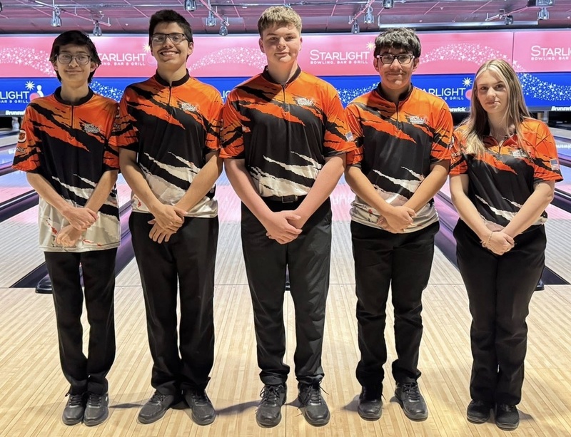 One female and four male teens in orange, black and white bowling shirts pose for a photo in front of bowling lanes.
