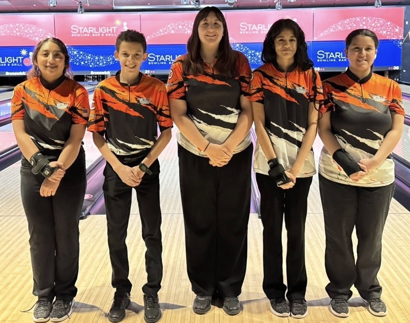 One male and four female teens in orange, black and white bowling shirts pose for a photo in front of bowling lanes.