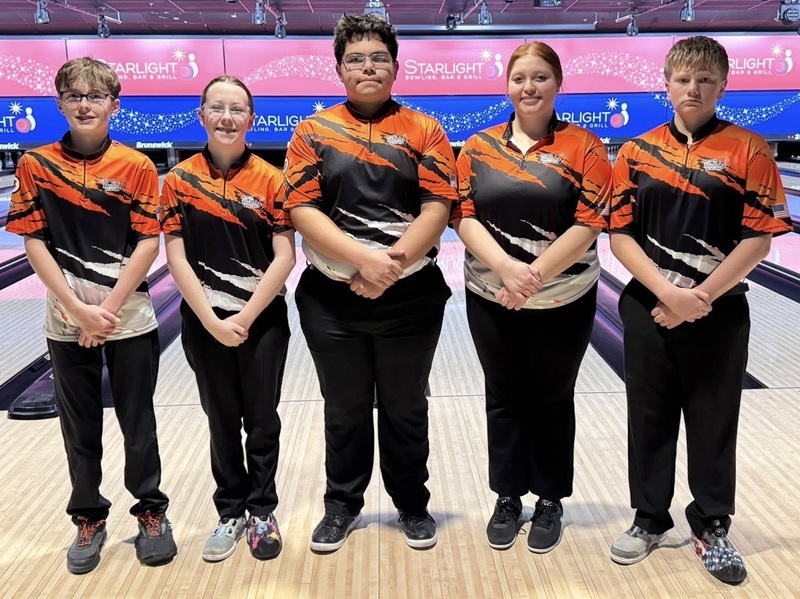Two female and three male teens in orange, black and white bowling shirts pose for a photo in front of bowling lanes.