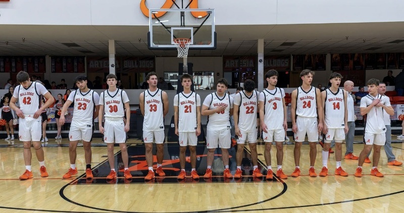 Eleven male teens in white and orange basketball uniforms and orange shoes stand in a straight line on a court awaiting the pregame introduction.