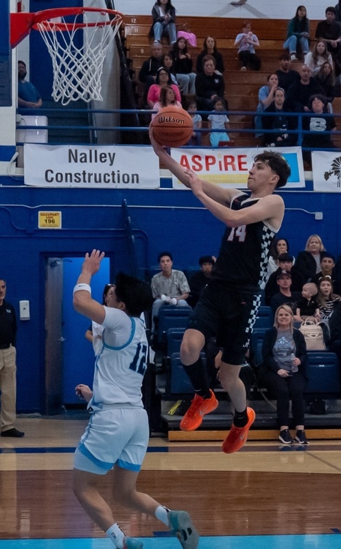 A teen boy in a black and white basketball uniform with orange shoes sails into the lane in mid-air with the ball extended in his right hand as an opponent in white and blue stands below him.