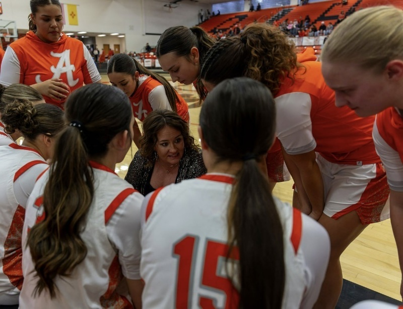 A group of teen girls in white and orange basketball uniforms gathers around a female coach who is kneeling on the court speaking to them.