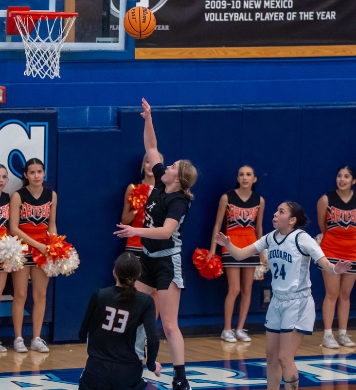 A teen girl in a black and white basketball uniform goes up under the basket and releases the ball from her right hand as a teammate and an opponent in white and blue look on.