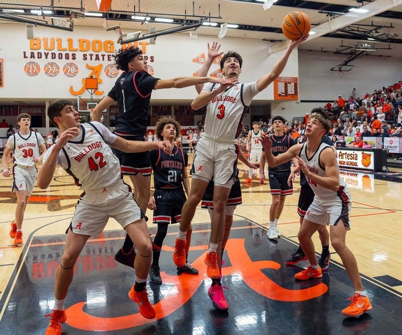 A teen boy in a white and orange basketball uniform goes toward the basket in mid-air with the ball extended in his left hand as two teammates and three opponents in black and red look on.
