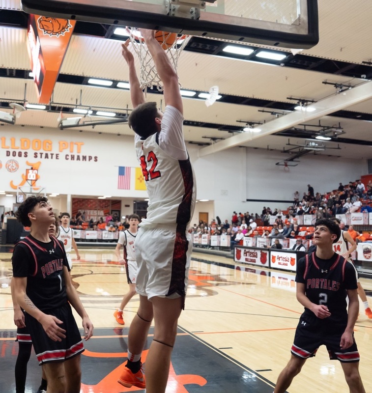 A teen boy in a white and orange basketball uniform dunks the ball as two opponents in black and red look on.
