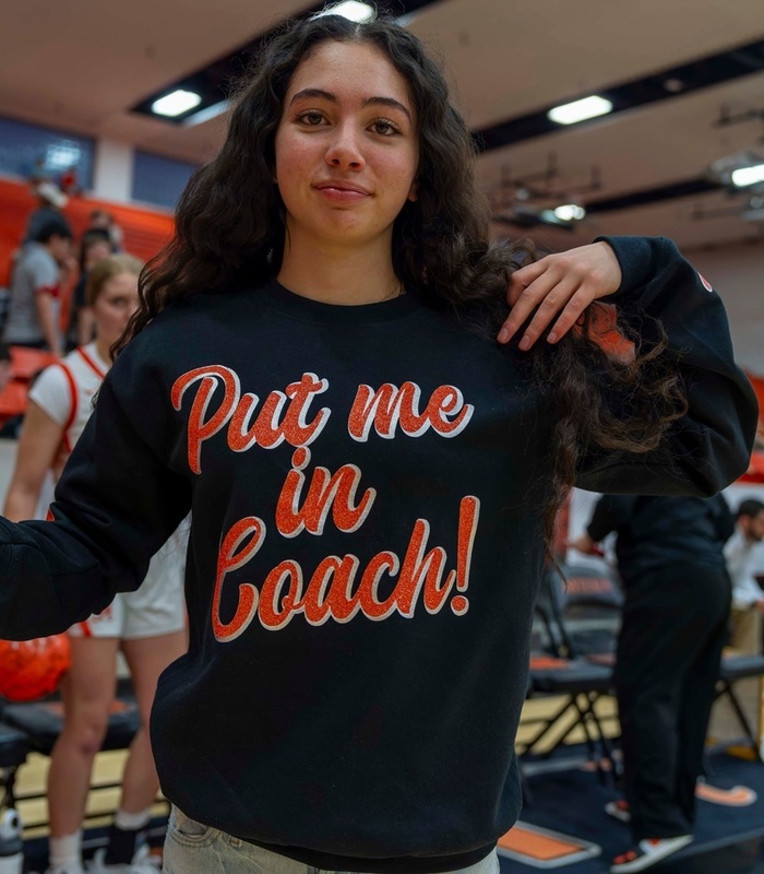 A teen girl poses for a photo wearing a black sweatshirt reading "Put me in Coach!" in orange lettering.