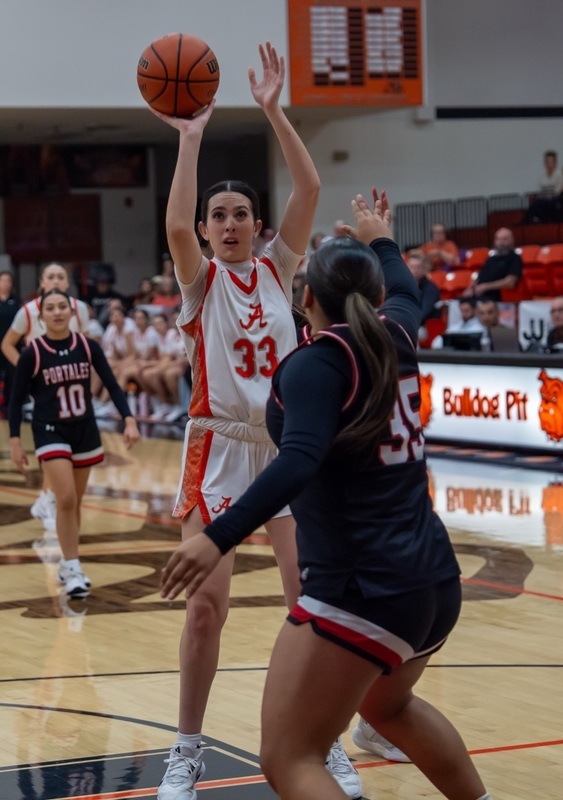 A teen girl in a white and orange basketball uniform prepares to release the ball from her right hand as an opponent in red and black moves in to defend.