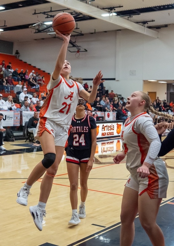 A teen girl in a white and orange basketball uniform jumps as she releases the ball from her right hand as a teammate and an opponent in black and red look on.