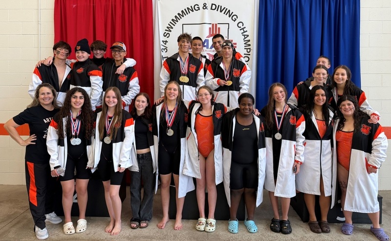 A large group of male and female teens in orange, black and white swimming warm-ups pose for a photo with a female coach. Many of the athletes have medals around their necks. 