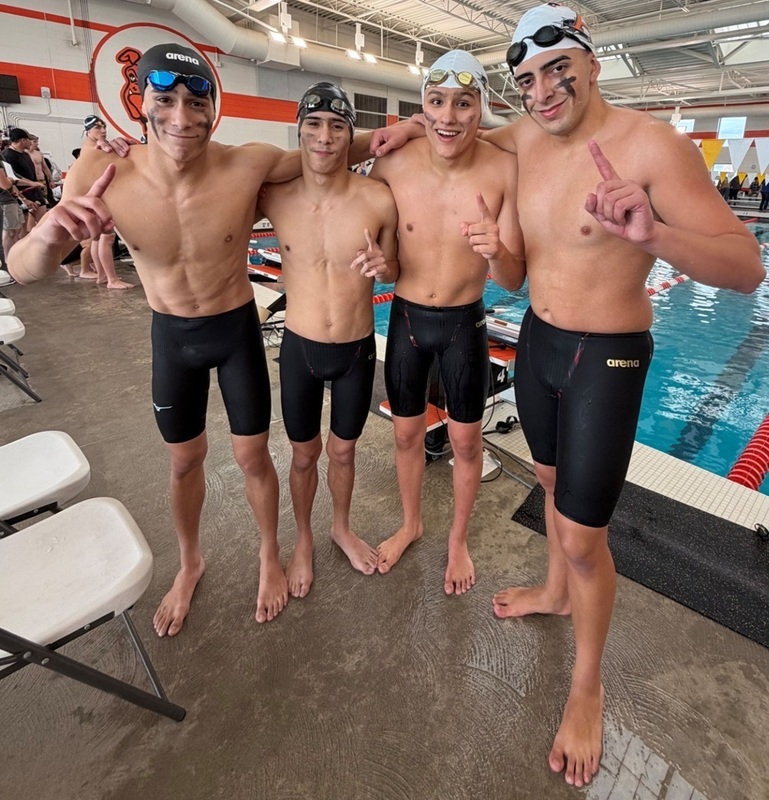 Four teen boys in swim trunks and swim caps pose for a photo alongside a pool holding their fingers up to indicate "number one".