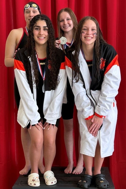 Four teen girls in orange, black and white swimming warm-ups pose for a photo atop a podium. They wear silver medals around their necks.