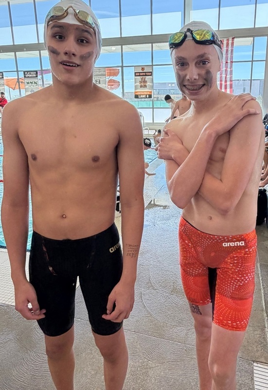 Two teen boys, one in black swim trunks and one in orange, both wearing swim caps and goggles pushed onto their foreheads, pose for a photo at a natatorium.