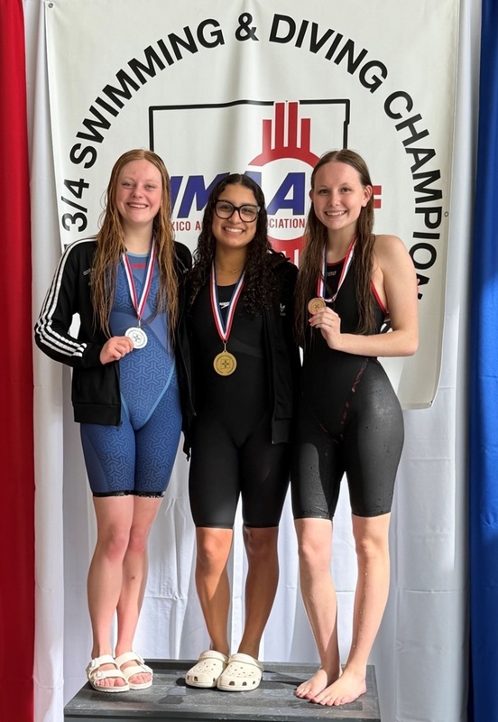 Three teen girls, one in a blue swimsuit and two in black, pose for a photo atop a podium wearing, from left, silver, gold and bronze medals around their necks.