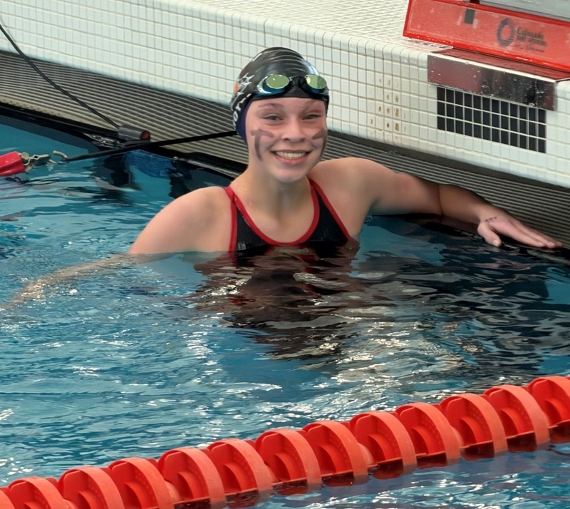 A teen girl in a black swimsuit and black swim cap with goggles pushed up onto her forehead smiles for the camera as she waits half-submerged in a swimming pool.