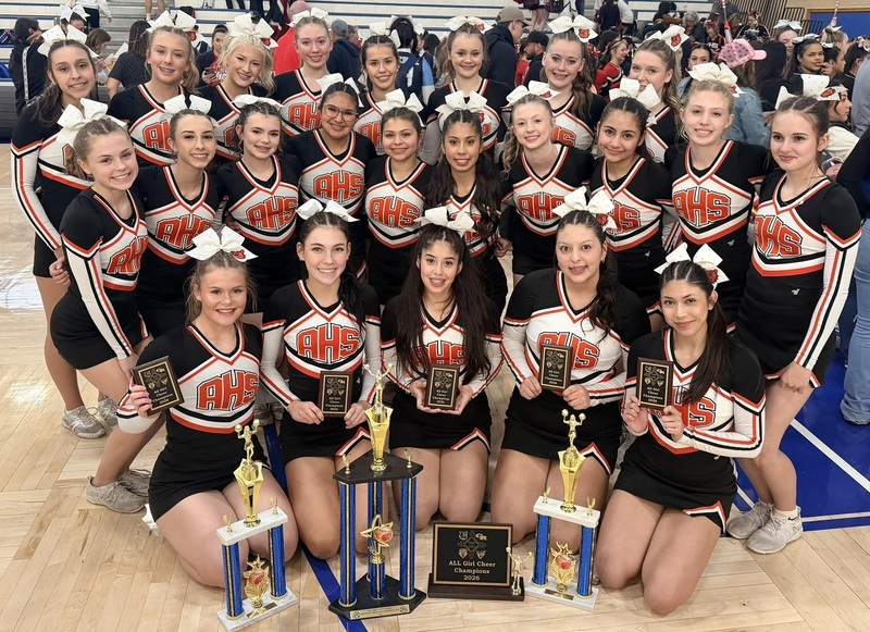 A large group of female teens in black, orange and white cheerleading uniforms with large white bows atop their heads poses for a photo. The front row holds small plaques and has four trophies placed in front of them.