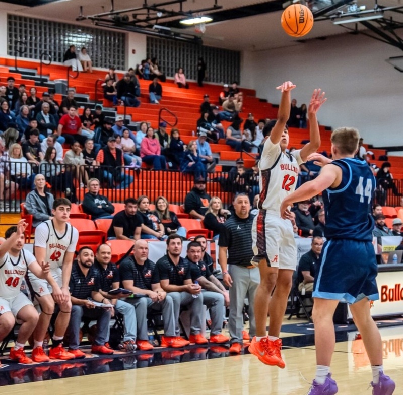 A teen boy in a white and orange basketball uniform jumps as he shoots the ball high into the air over a sizable defender in blue. Behind him, his team's bench and a portion of the crowd look on in anticipation.
