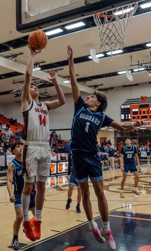A teen boy in a white and orange basketball uniform hops under the basket as he releases the ball from his right hand. An opponent in blue jumps in front of him with his right arm up to defend.