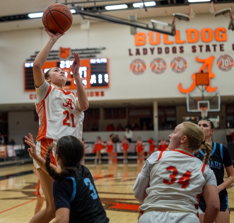 A teen girl in a white and orange basketball uniform is seen from the knees up as she lifts the ball with her right hand to release it. A teammate and two opponents in blue are visible below her.