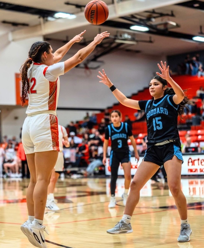 A teen girl in a white and orange basketball uniform goes straight up into the air as she releases the ball toward the basket. An opponent in blue raises both arms in an attempt to defend.