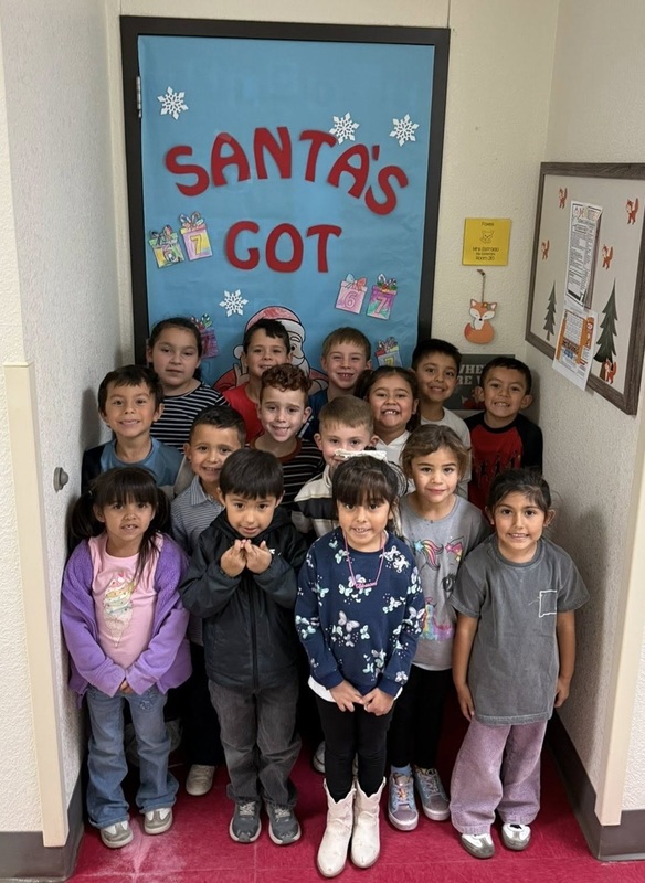 A group of male and female children pose for a photo in front of a classroom door that reads "Santa's Got Gifts for Us".