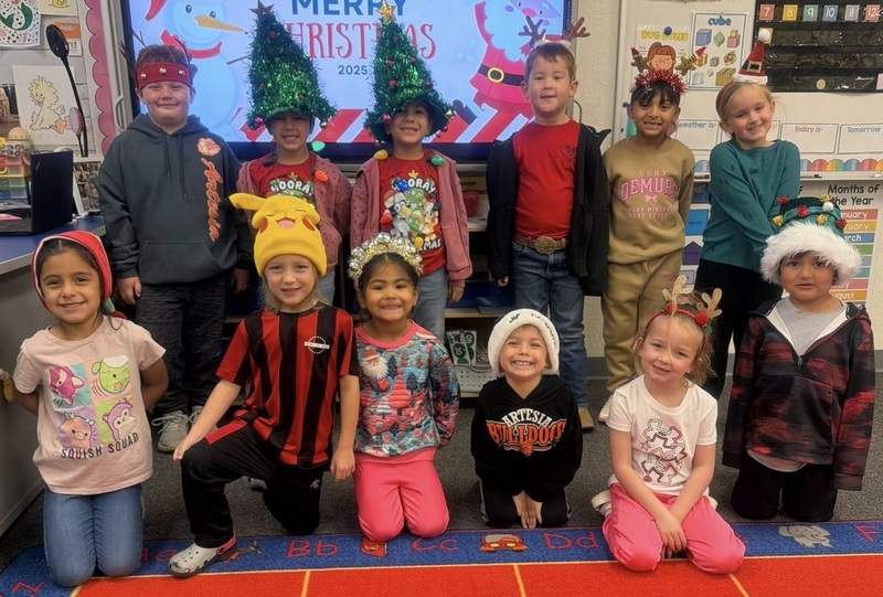 A group of male and female children pose for a photo. The children wear different holiday hats on their heads, including two with trees made of tinsel.