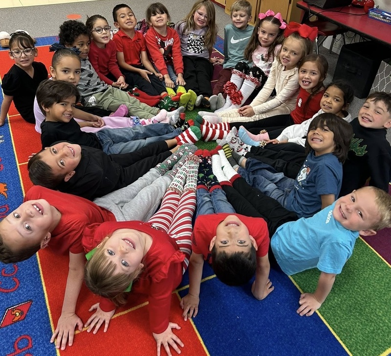 A group of male and female children smile for the camera while sitting in a circle with their feet together in the middle. The children wear brightly decorated Christmas socks.