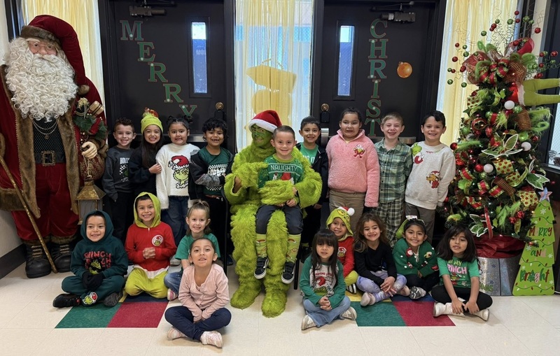 A group of male and female children pose for a photo gathered around a person dressed as the Grinch. The Grinch holds one child and points to her shirt, which reads "Naughty".
