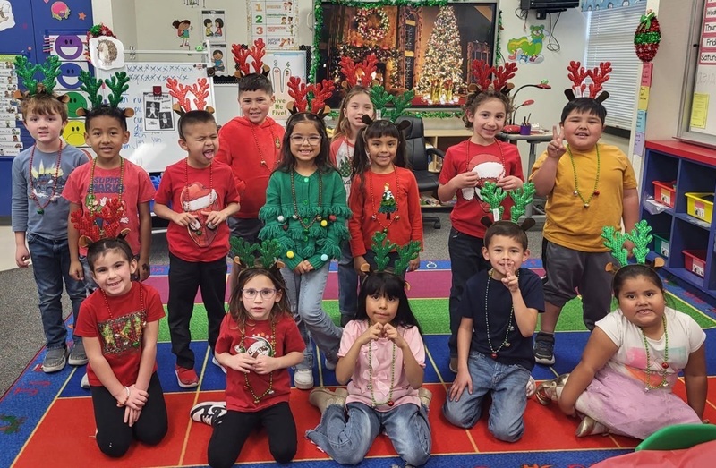 A group of male and female children pose for a photo. The children wear brightly colored Christmas shirts and many also wear reindeer headbands.