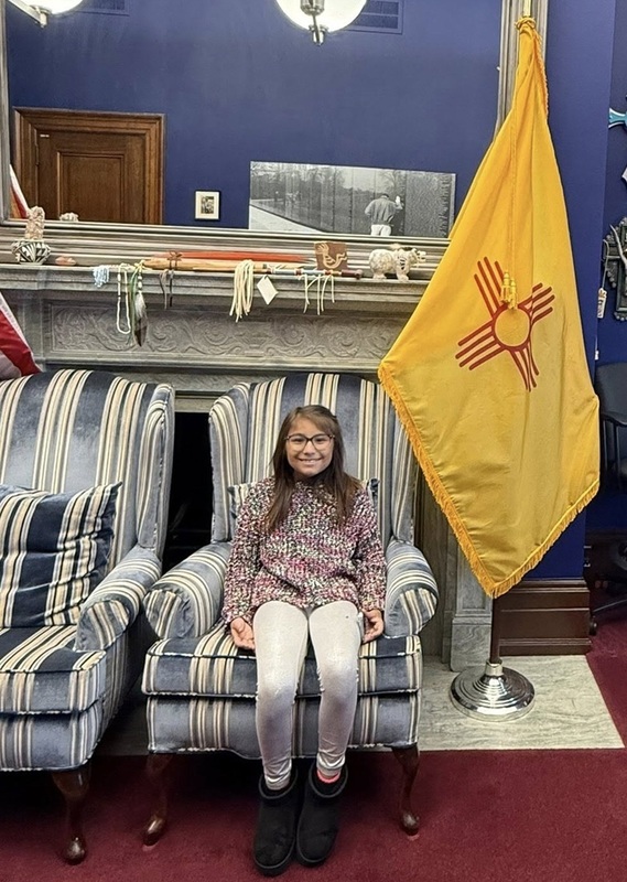 A female child sits in a chair adjacent a New Mexico flag in the White House.